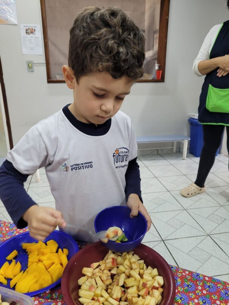 Criança do Colégio Futura Jundiaí se servindo de salada de frutas com manga e maçã em vivência de alimentação saudável.