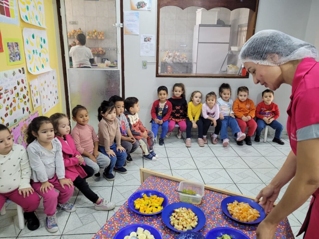 Nutricionista Ana Paula Silva com touca higiênica e jaleco rosa mostrando pratos de frutas picadas para crianças atentas do Colégio Futura Jundiaí.