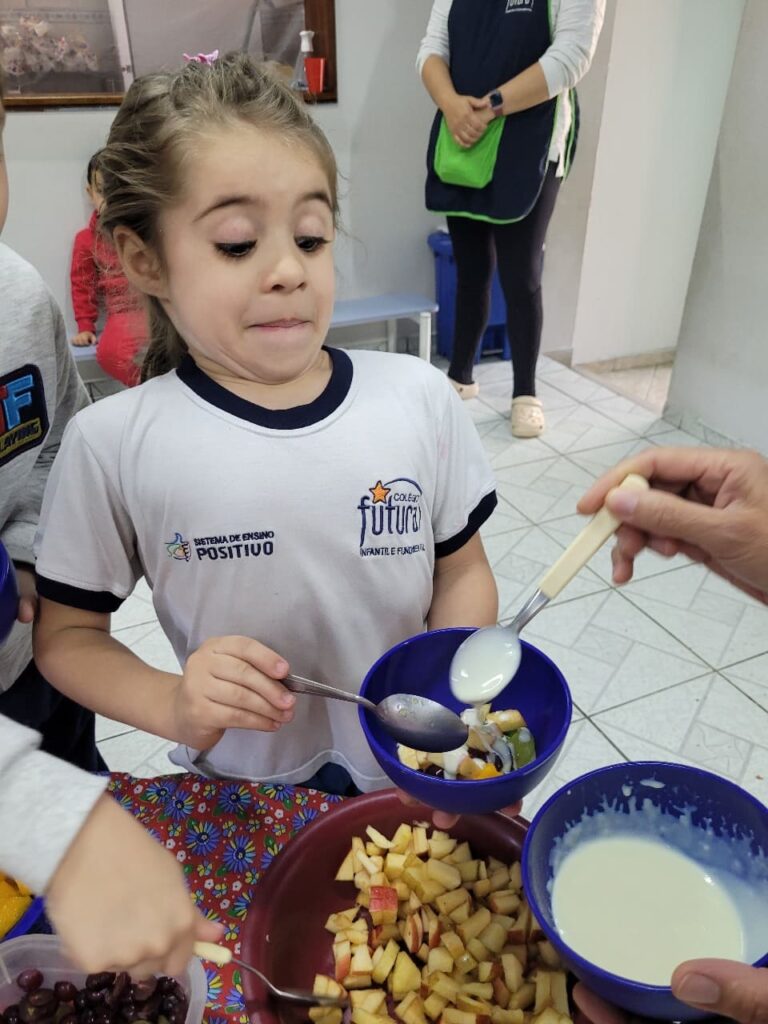 Criança do Colégio Futura Jundiaí com expressão curiosa adicionando iogurte na salada de frutas em vivência sensorial.