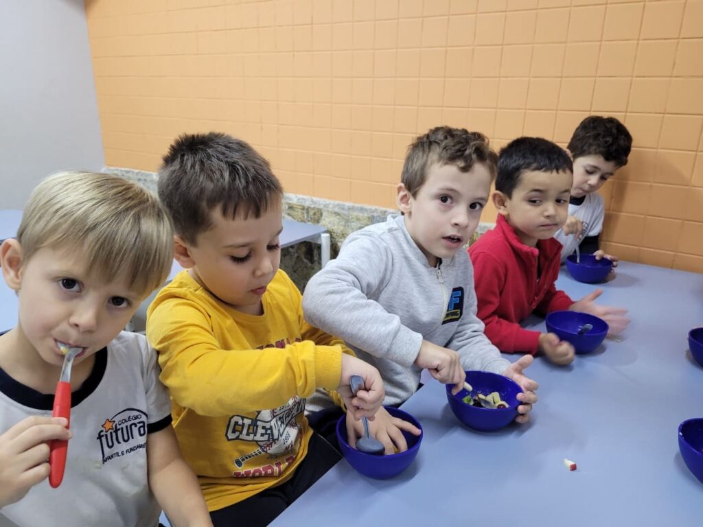 Grupo de crianças do Colégio Futura Jundiaí comendo salada de frutas com colheres em aula de alimentação saudável.