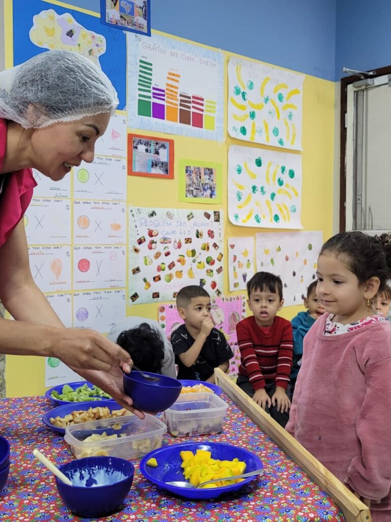 Nutricionista servindo manga para criança em tigela azul durante vivência de frutas no Colégio Futura Jundiaí.