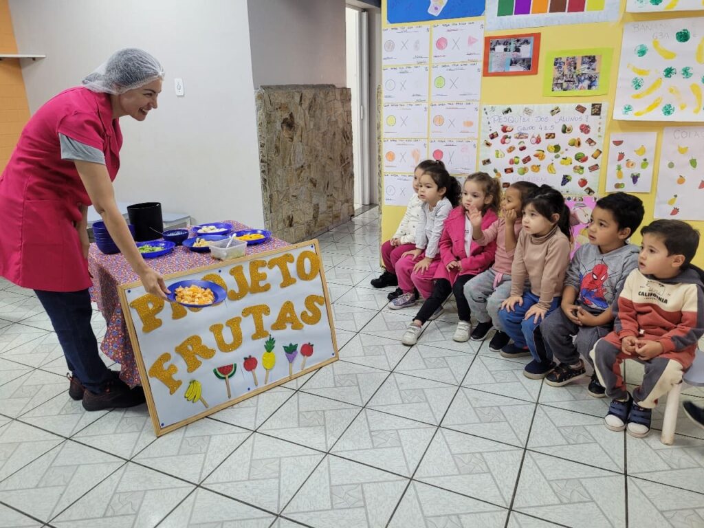 Nutricionista e crianças do Colégio Futura Jundiaí ao lado do painel Projeto Frutas, prestes a iniciar a degustação.