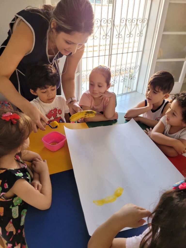 Professor e crianças do Colégio Futura Jundiaí usando banana para registrar a cor amarela em papel durante vivência sensorial com frutas.