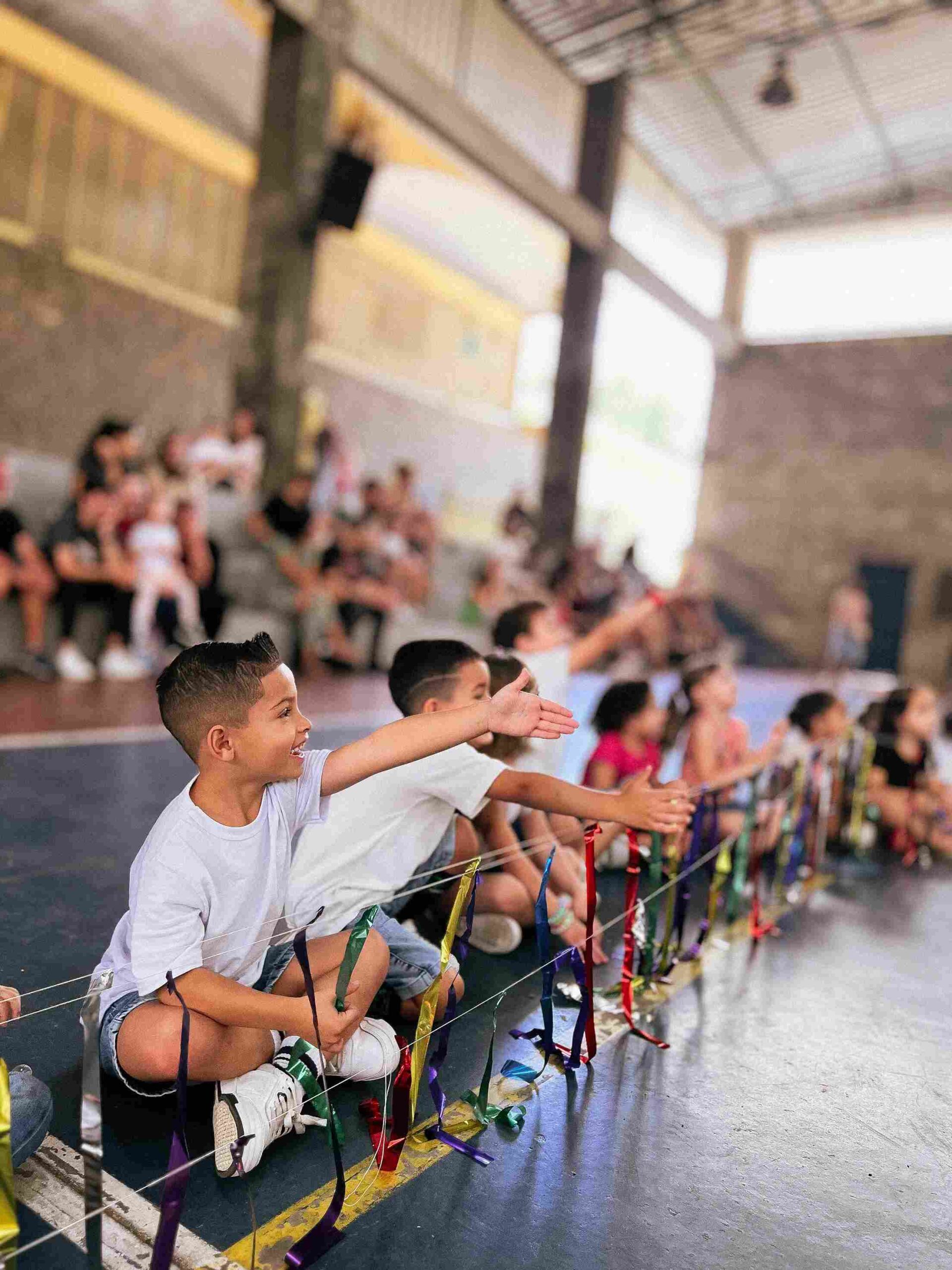 Crianças da plateia sorrindo e estendendo a mão para as fitas coloridas durante o Sarau 2025 do Colégio Futura Infantil Jundiaí.