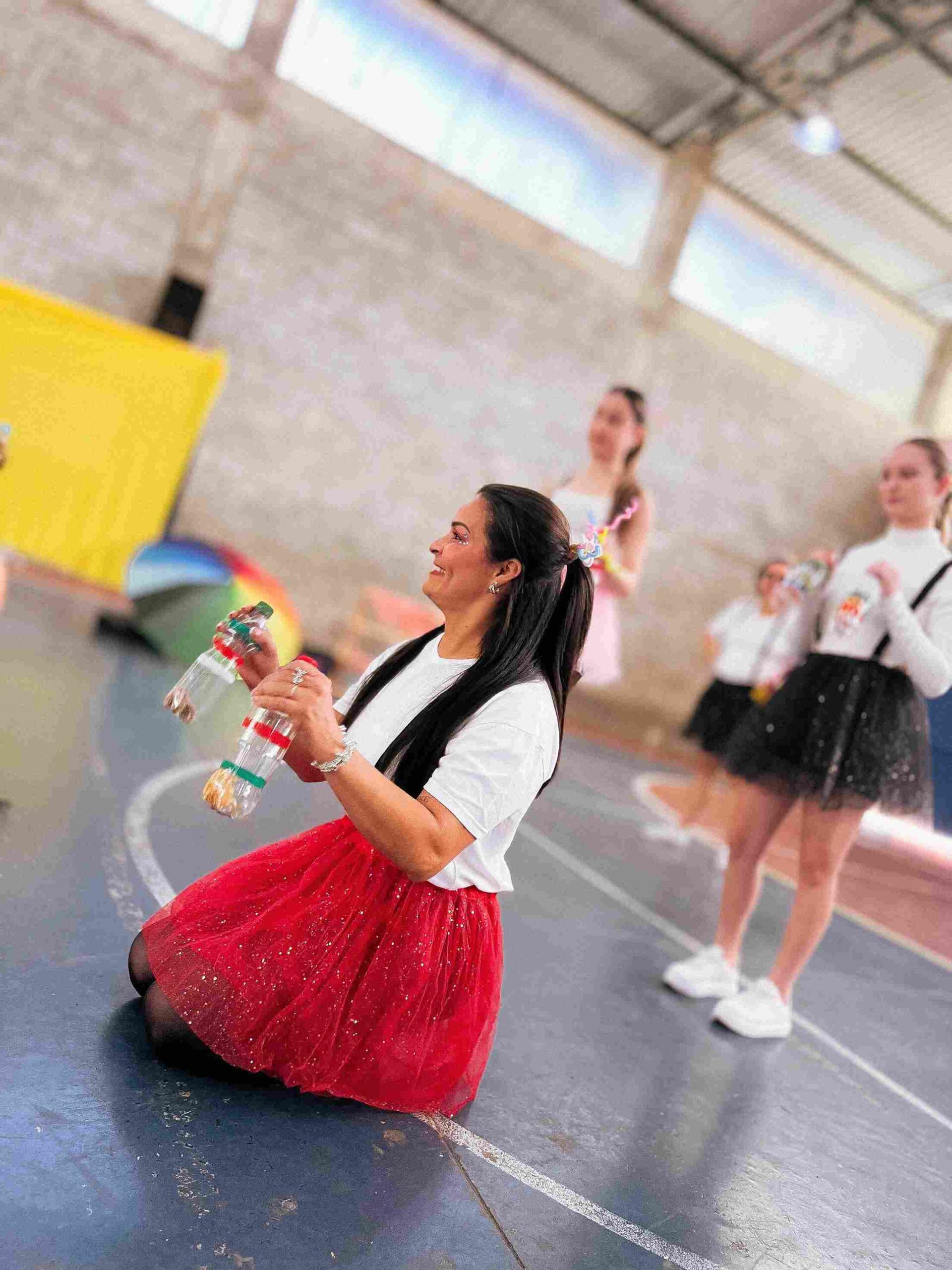 Professora do Colégio Futura Jundiaí ajoelhada, sorrindo e usando garrafas PET como instrumento musical no Sarau 2025.