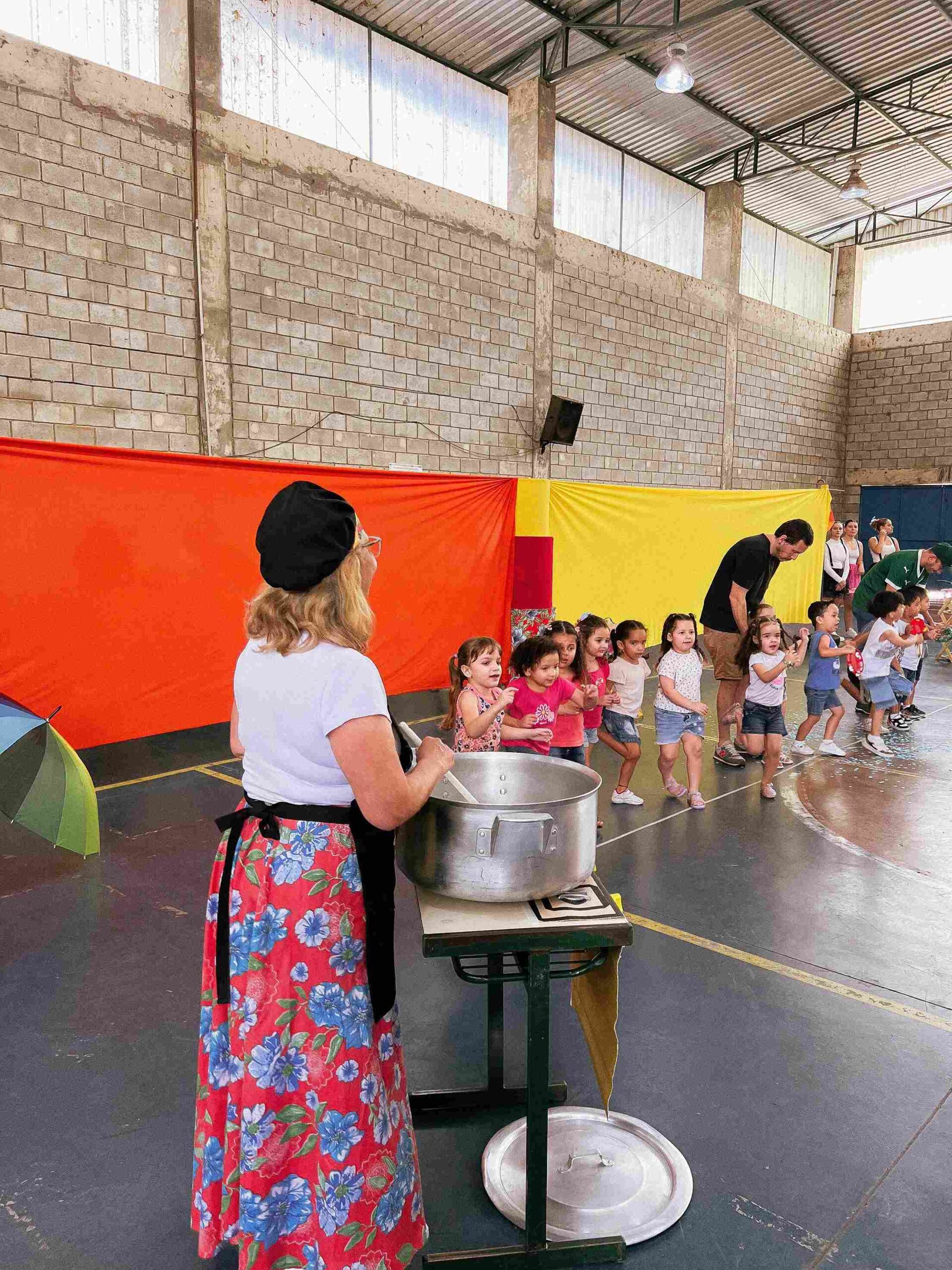 Professora com caldeirão gigante cozinhando e crianças se aproximando na apresentação do Sarau 2025 do Colégio Futura Jundiaí.